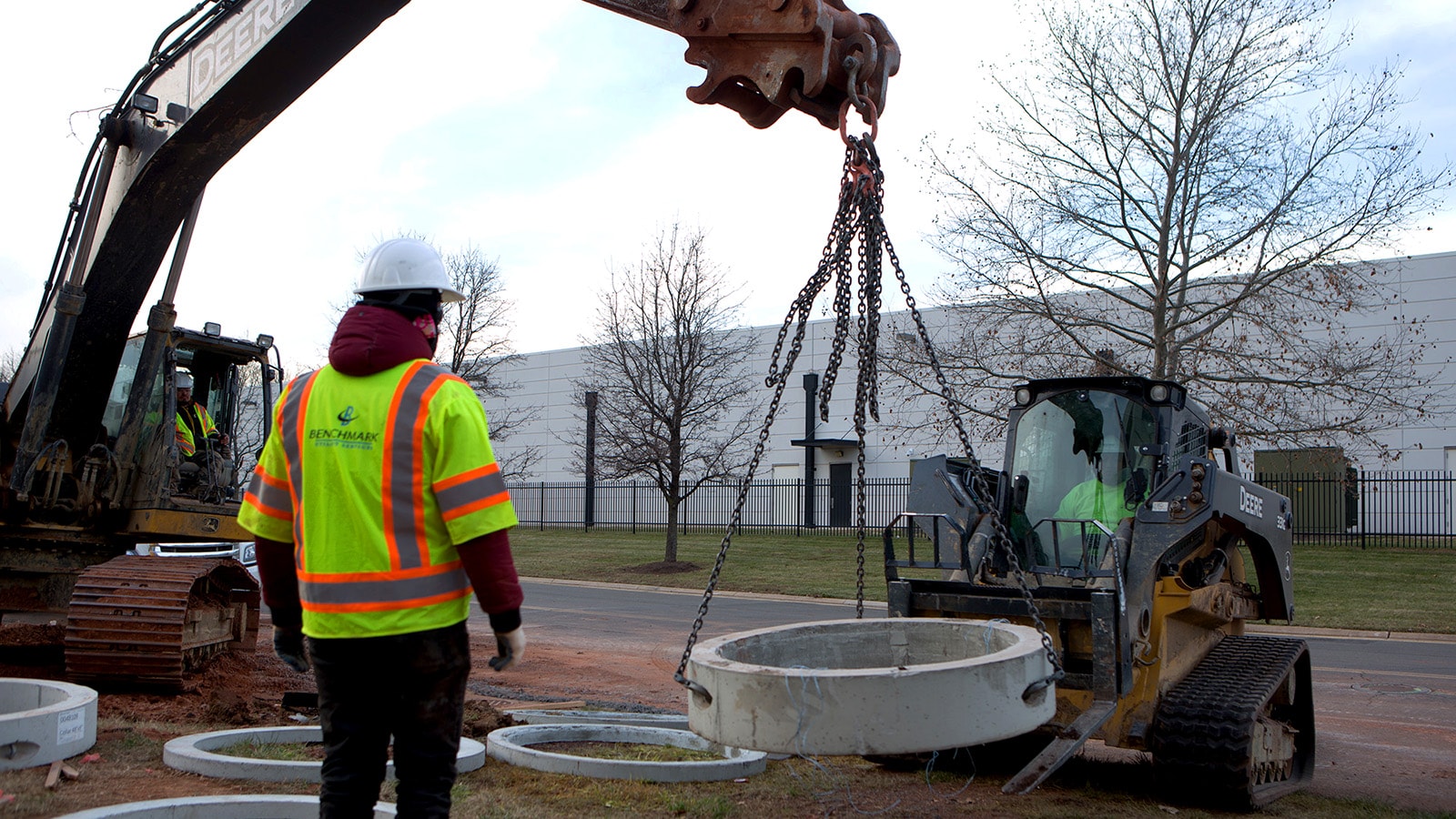 An excavator lifts a section of concrete pipe off a compact track loader’s forks while a worker supervises nearby.