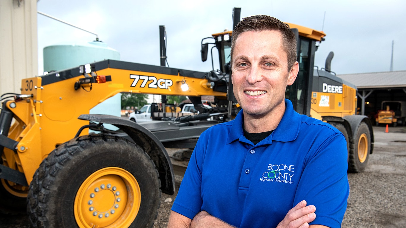 Nick Parr stands in front of a 772GP Motor Grader