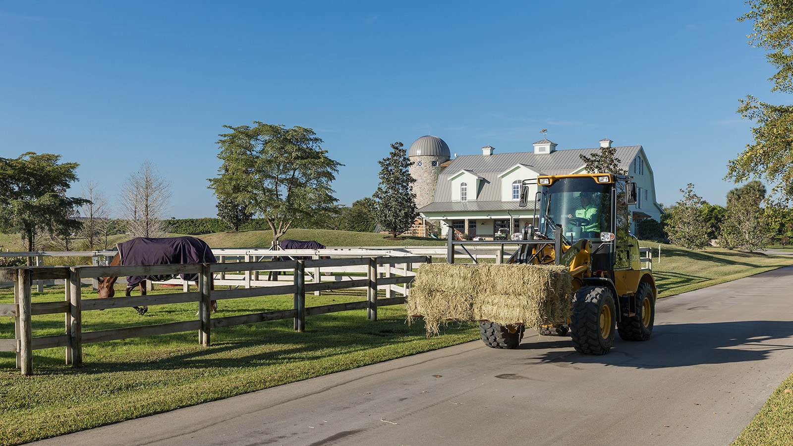Compact wheel loader hauls a bale of straw past a horse in a fenced area