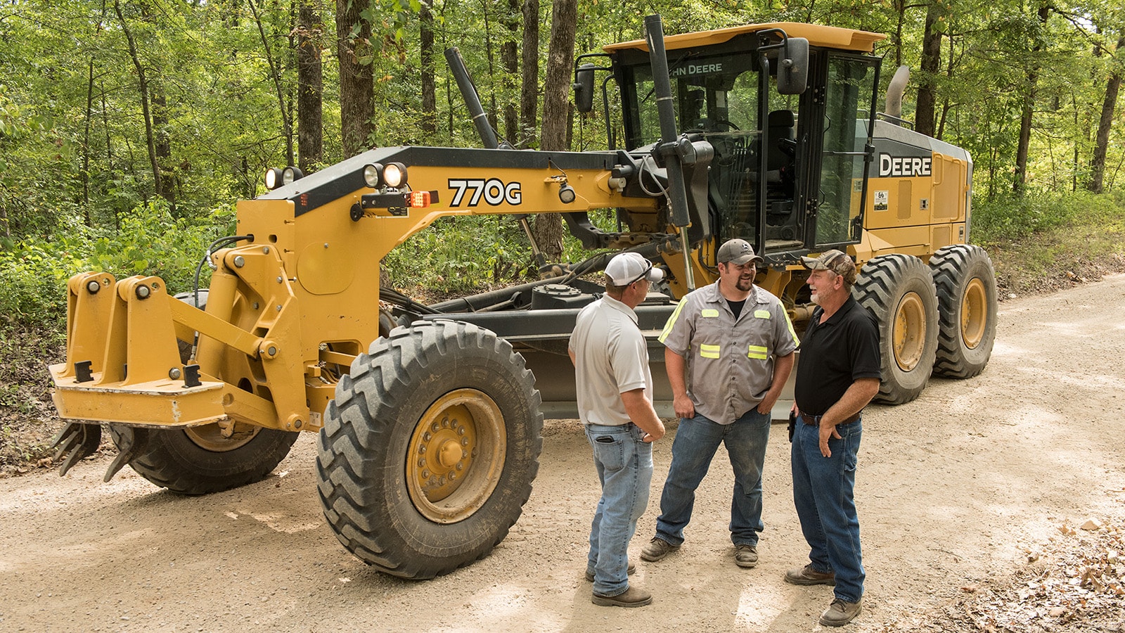 Three men stand in front of a 770G motor grader talking on a wooded gravel road