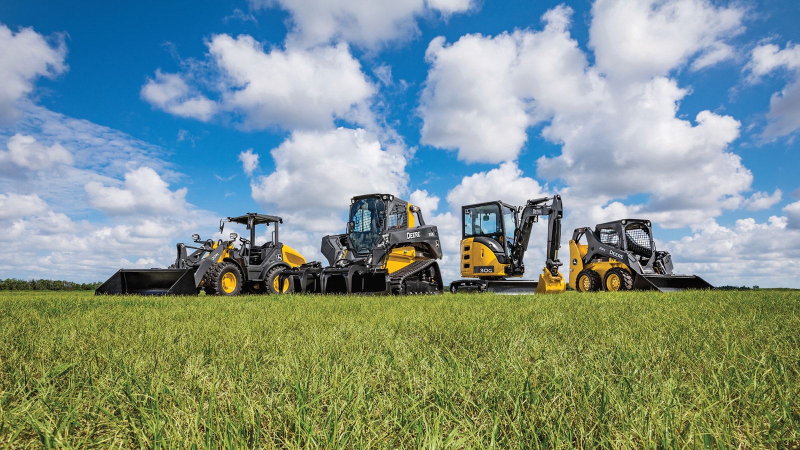 204L compact loader, 333G tracked skid steer, 30g mini excavator and 314G wheeled skid steer parked in a green grassy field with blue sky with white clouds in the background
