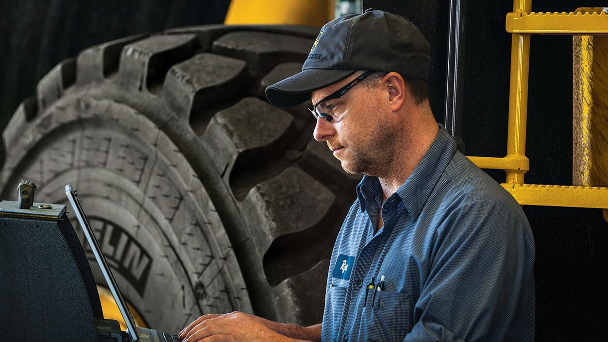 A technician types on a laptop in front of a piece of large construction equipment.