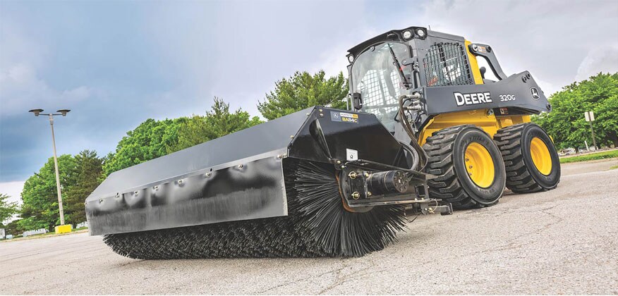 Una Carretilla Elevadora 320G de John Deere barre una playa de estacionamiento con un accesorio de Barredora Recolectora BA84C