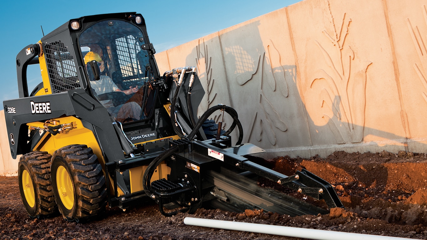 John Deere skid steer with trencher attachment digging a trench next to a concrete wall.