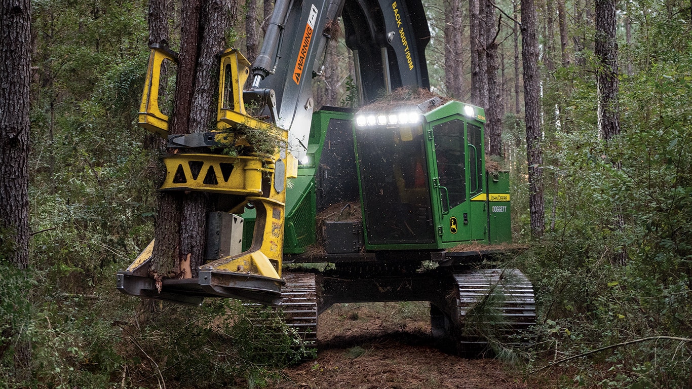 A John Deere 853M Tracked Feller Buncher with FR50 felling head is felling a tree in the forest.