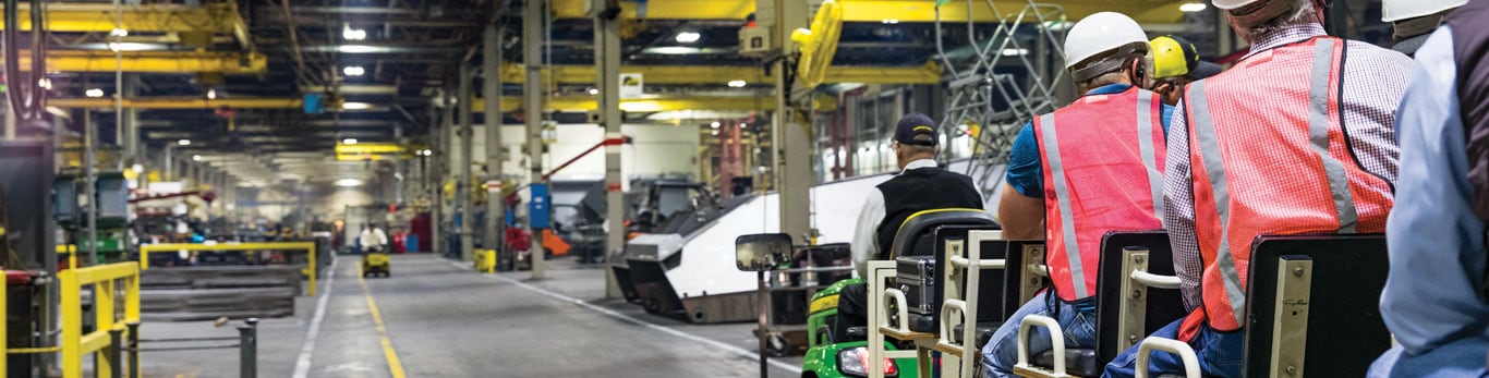 Tour participants wearing hardhats and riding on a trailer through a factory
