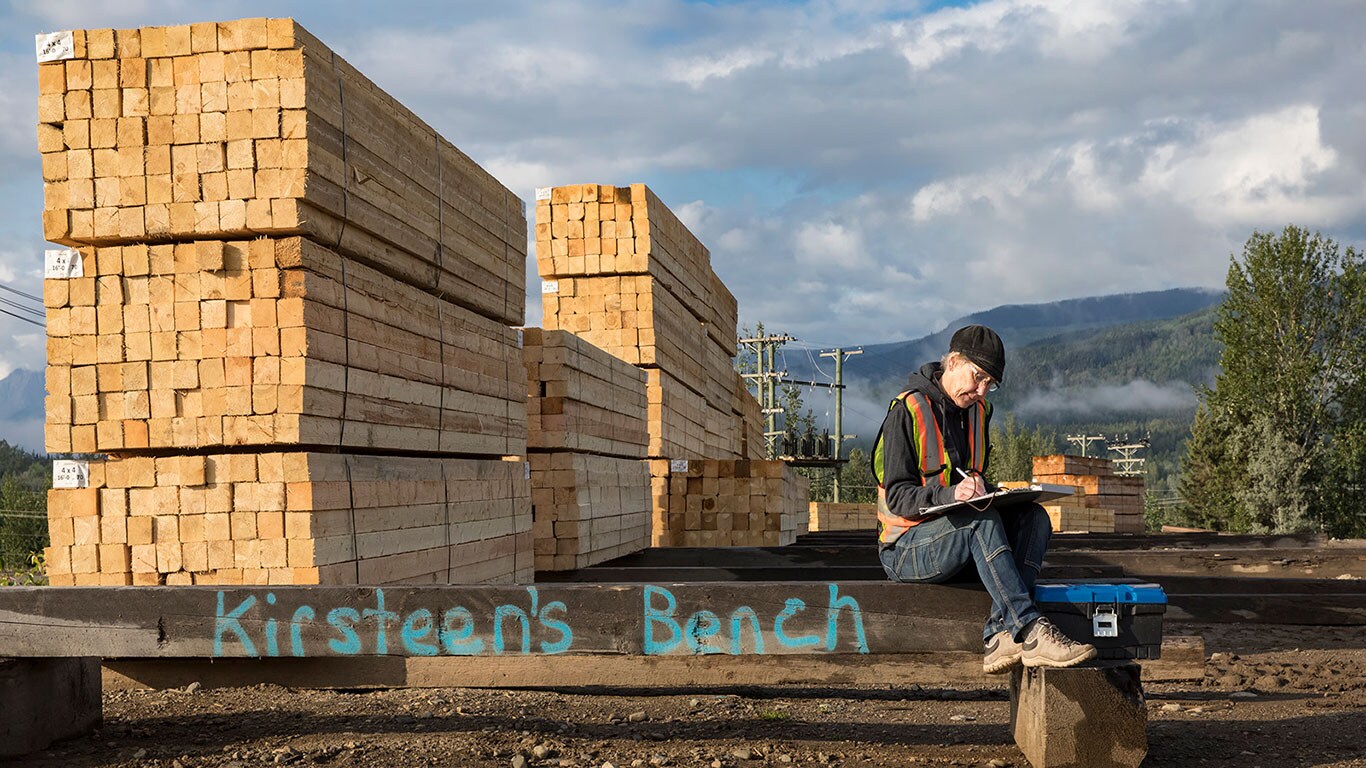 Kirsteen Laing working off a clipboard while sitting on a homemade bench next to stacks of lumber.
