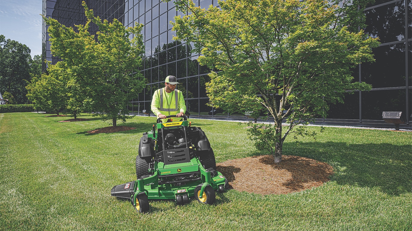 Outdoor photo of a professional landscape contractor mowing the grass around a young tree surrounded by dirt and mulch with the background of a tall, wide black corporate building that has dark windows.