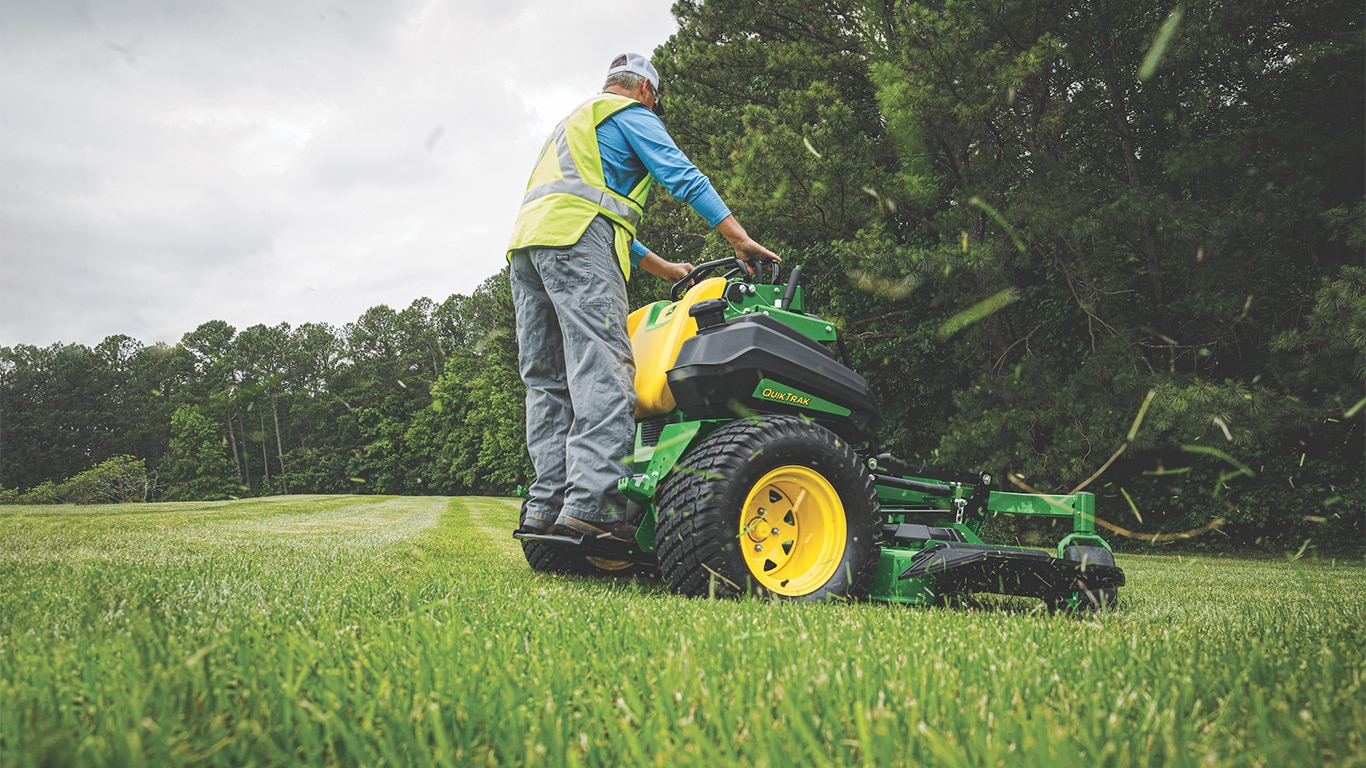 Outdoor picture with the camera from the ground angle looking up at a professional landscape contractor operation one of the new John&nbsp;Deere QuikTrak 800 Series Stand-On Commercial Mowers.