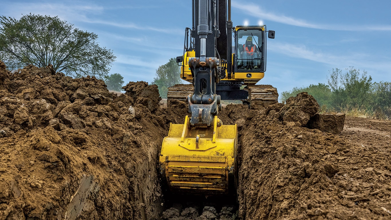 A yellow John Deere excavator uses its arm and bucket to dig a large hole at a construction site in a dirt field.