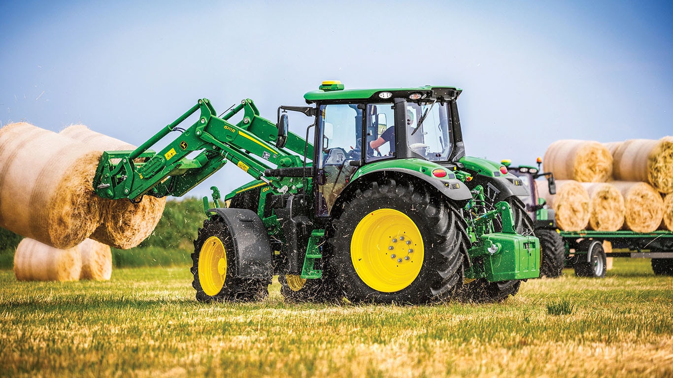 6M Tractor with 623R Loaders and bale spear with a round bale being transported in a field on a sunny day with clouds in the sky