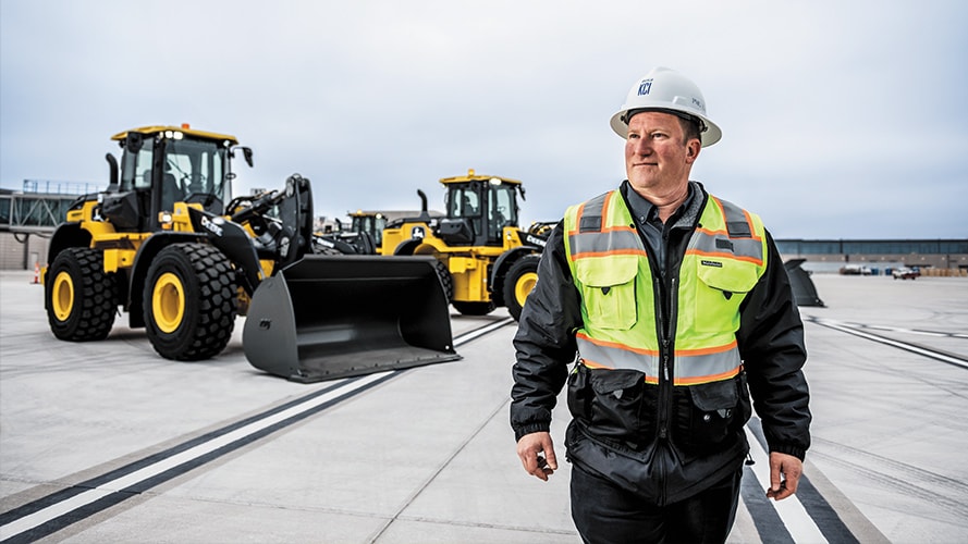 A construction worker walking onsite with Deere construction in the background