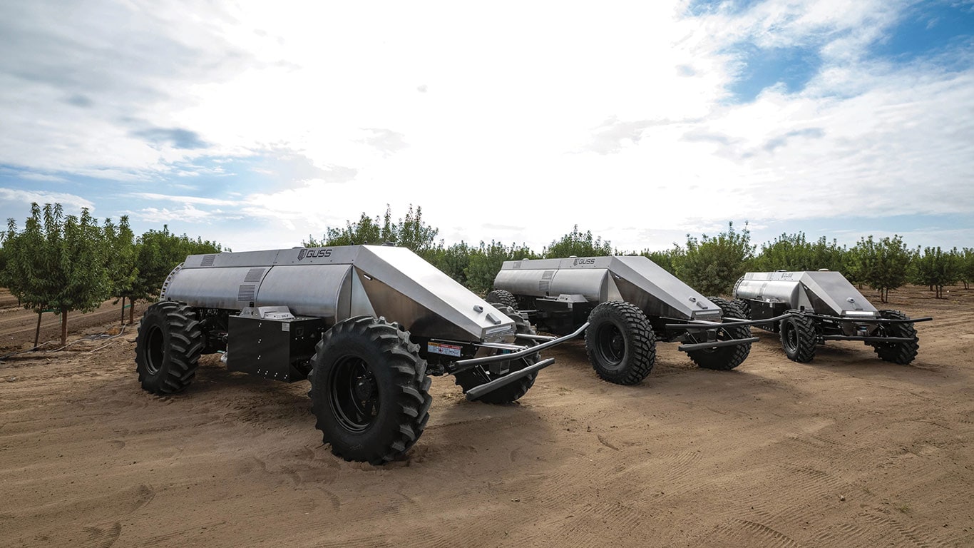 Three Guss Automation sprayers lined up in front of field of trees.