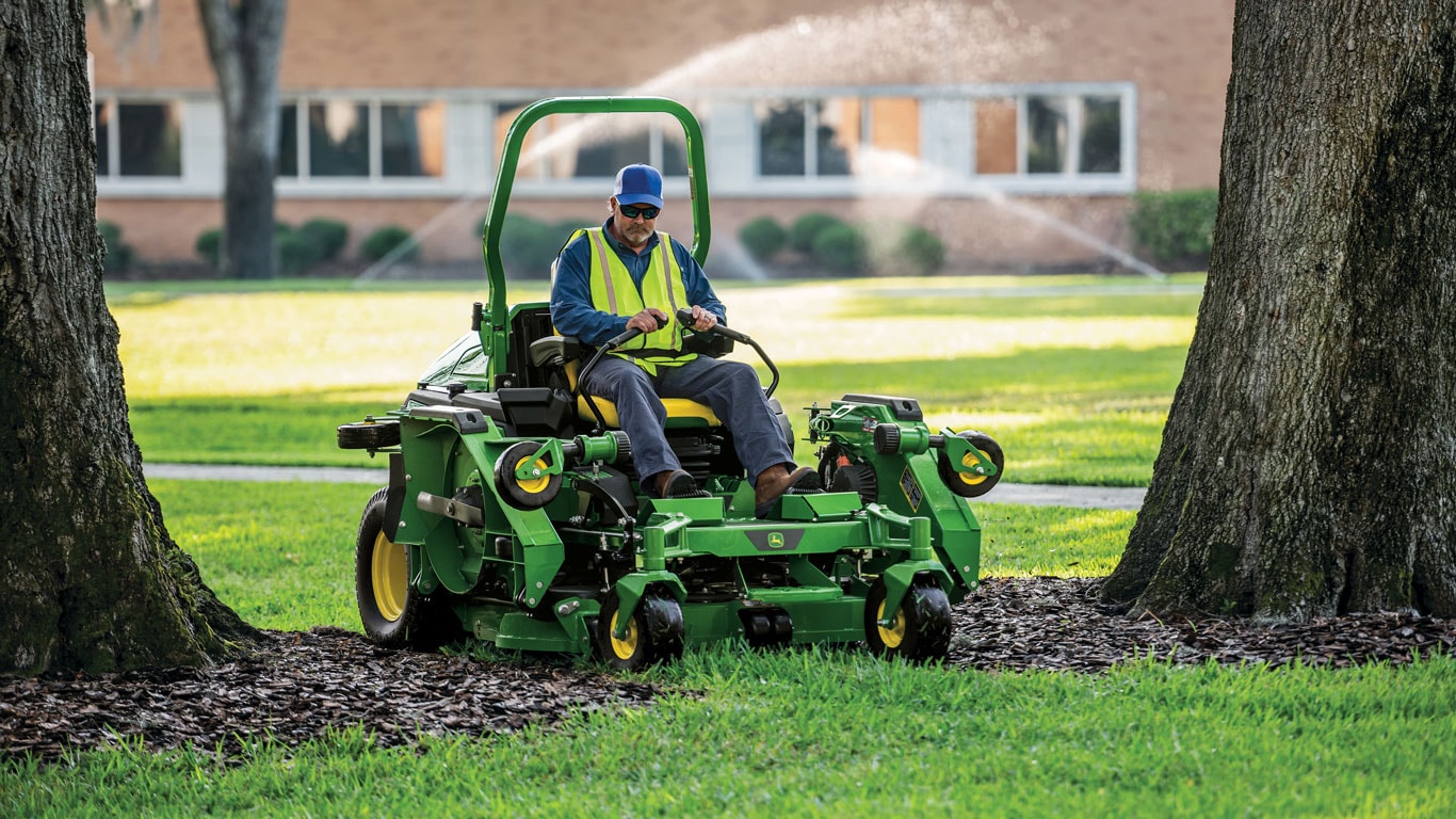 Operator on a John Deere ZTrak mowing in-between two trees with sprinklers watering lawn in the far background