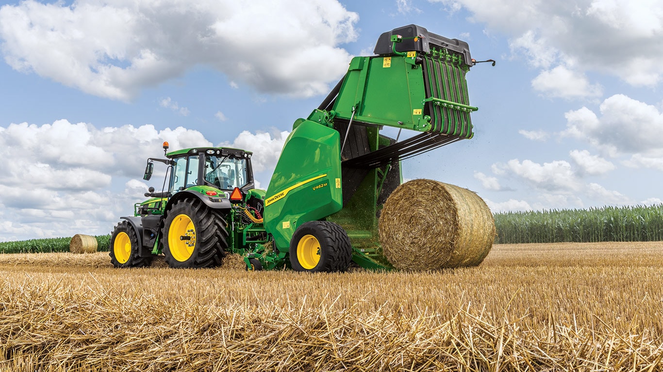 New John Deere V462M Round Baler by a John Deere 6M Tractor on a beautiful day with clouds in the sky and the baler is kicking out a round bale with the baler gate open
