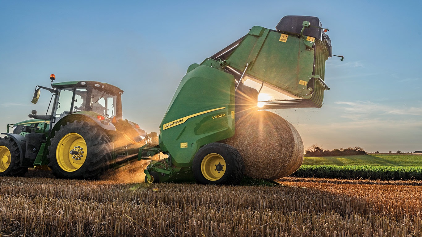 New John Deere V462M Round Baler being pulled by John Deere 6M Tractor with the baler gate open and a round bale being ejected as the sun peeks through the side of the baler during sunset