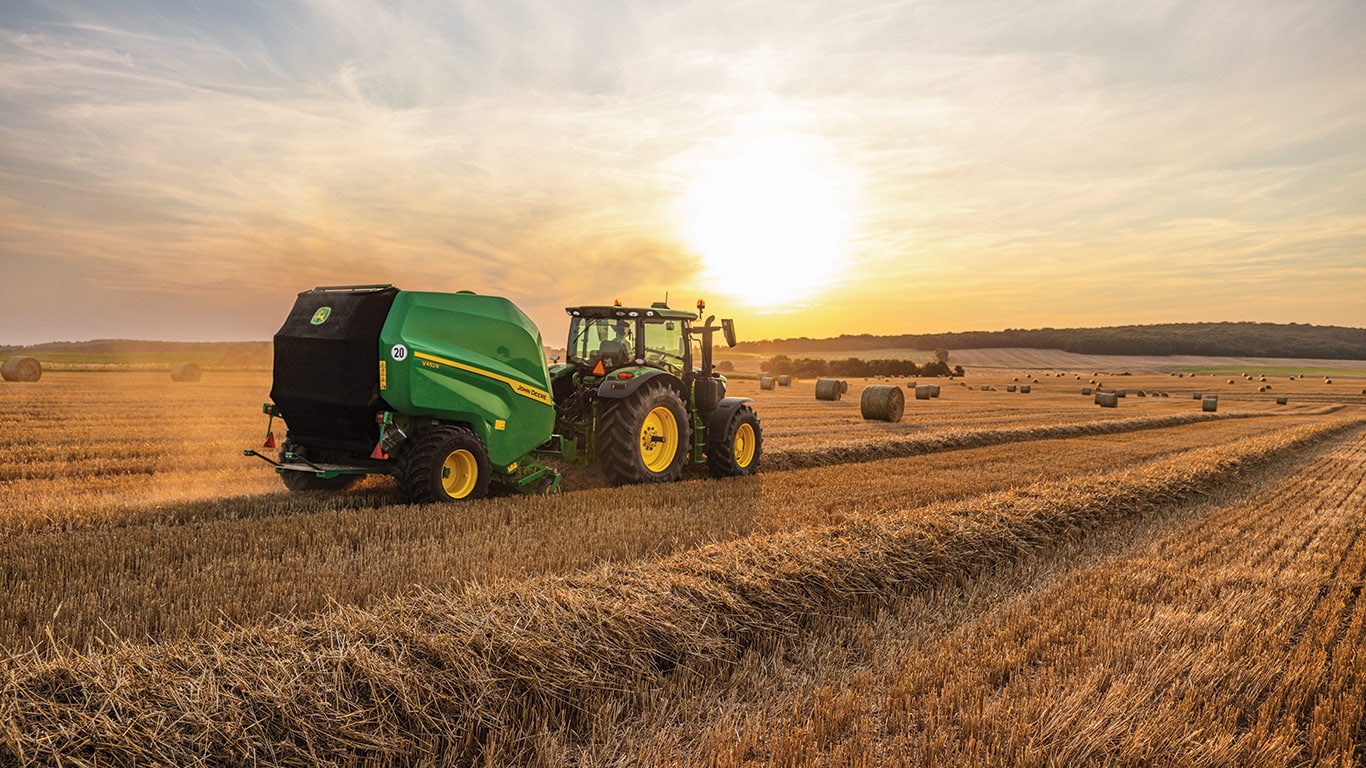New John Deere V462M Round Baler being pulled by a John Deere Tractor at dusk in a wide open field with dozens of round bales sitting in the field