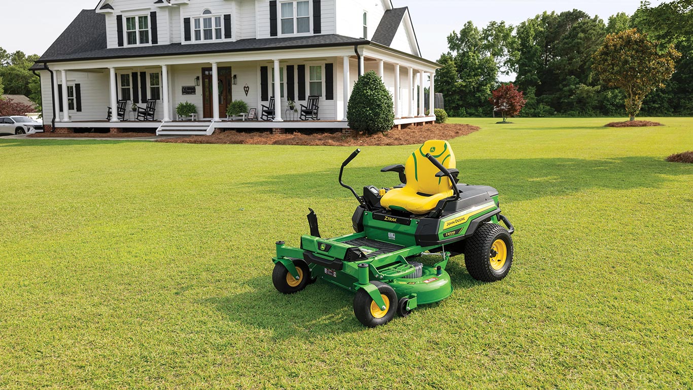 New John Deere Z370RS Electric ZTrak Mower featuring EGO 56V ARC Lithium™ Batteries parked on the front lawn of a large property with a beautiful white home in the background with an expansive deck an deck chairs for to relax after mowing is done.