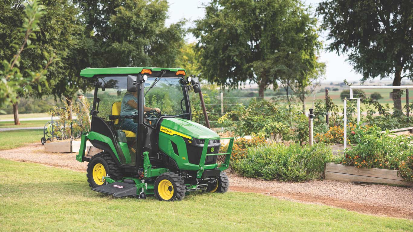 Green compact tractor mowing grass beside garden beds and a gravel path in a landscaped area.