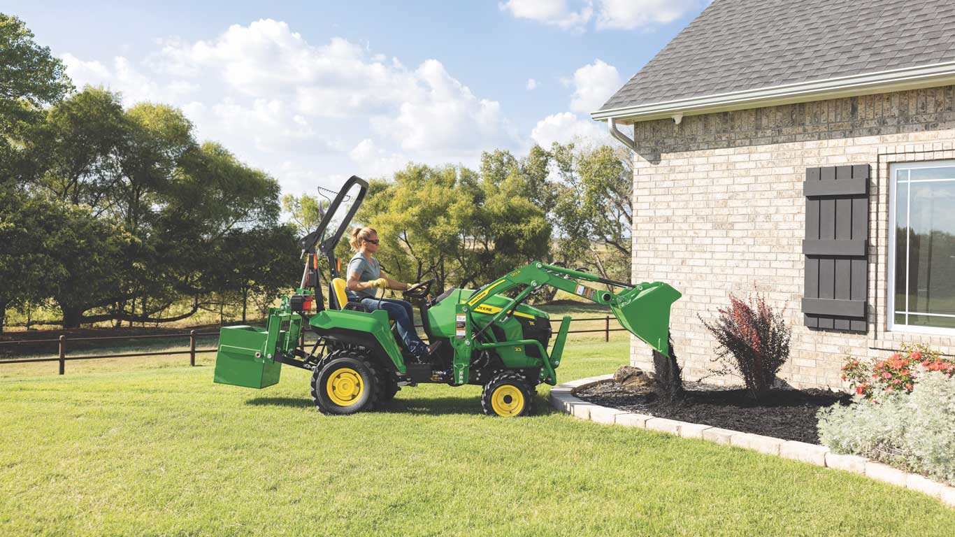 Compact tractor with front loader lifting soil from a landscaped bed beside a brick house.