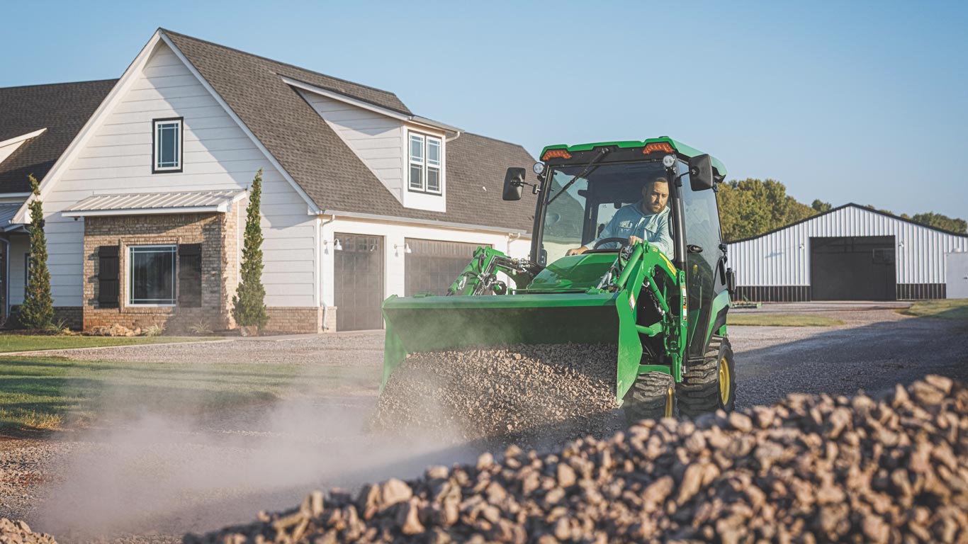 Compact tractor spreads gravel on a driveway in front of a rural home and barn.