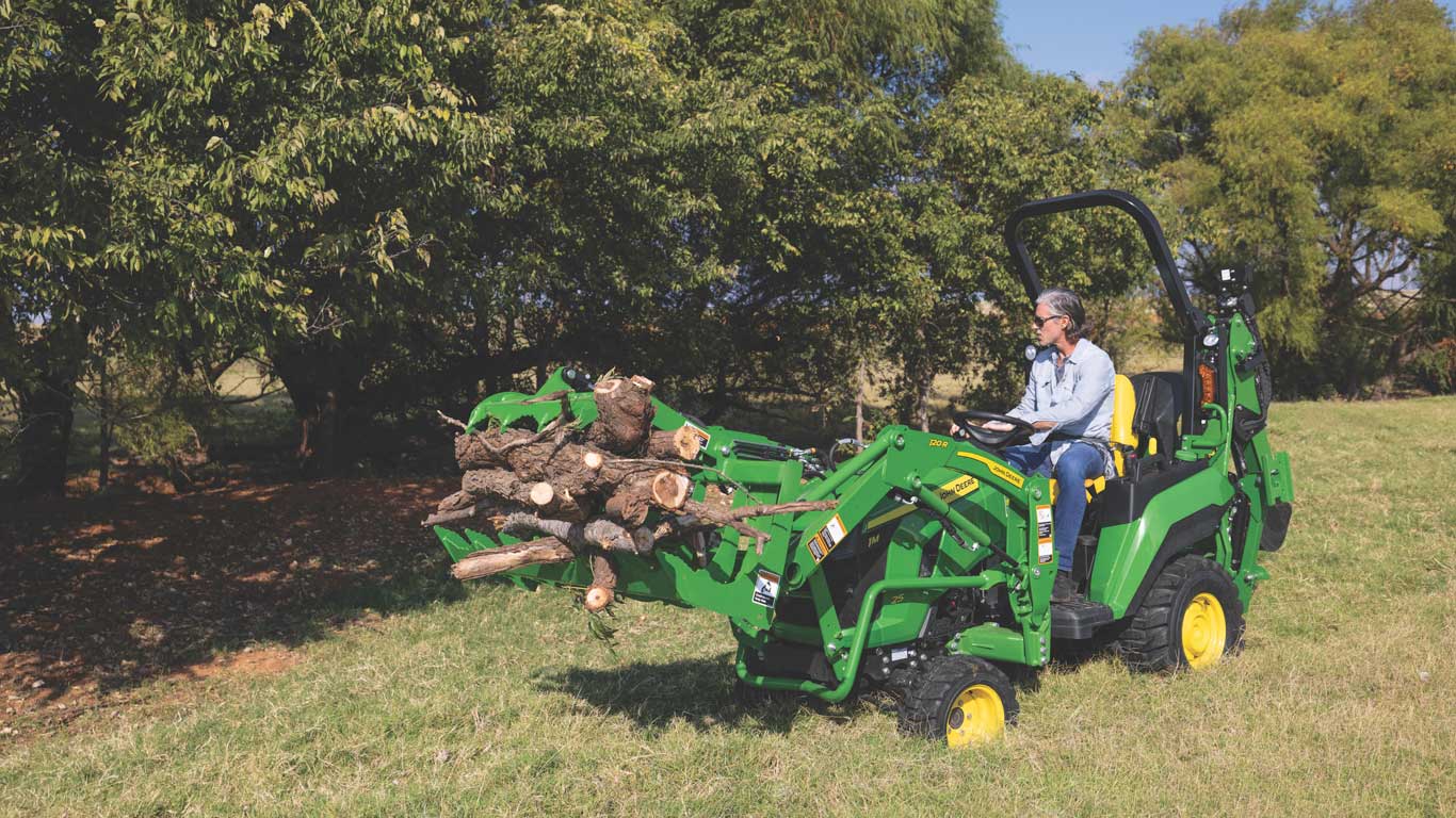 Compact tractor moves a load of cut logs across a grassy field near trees.