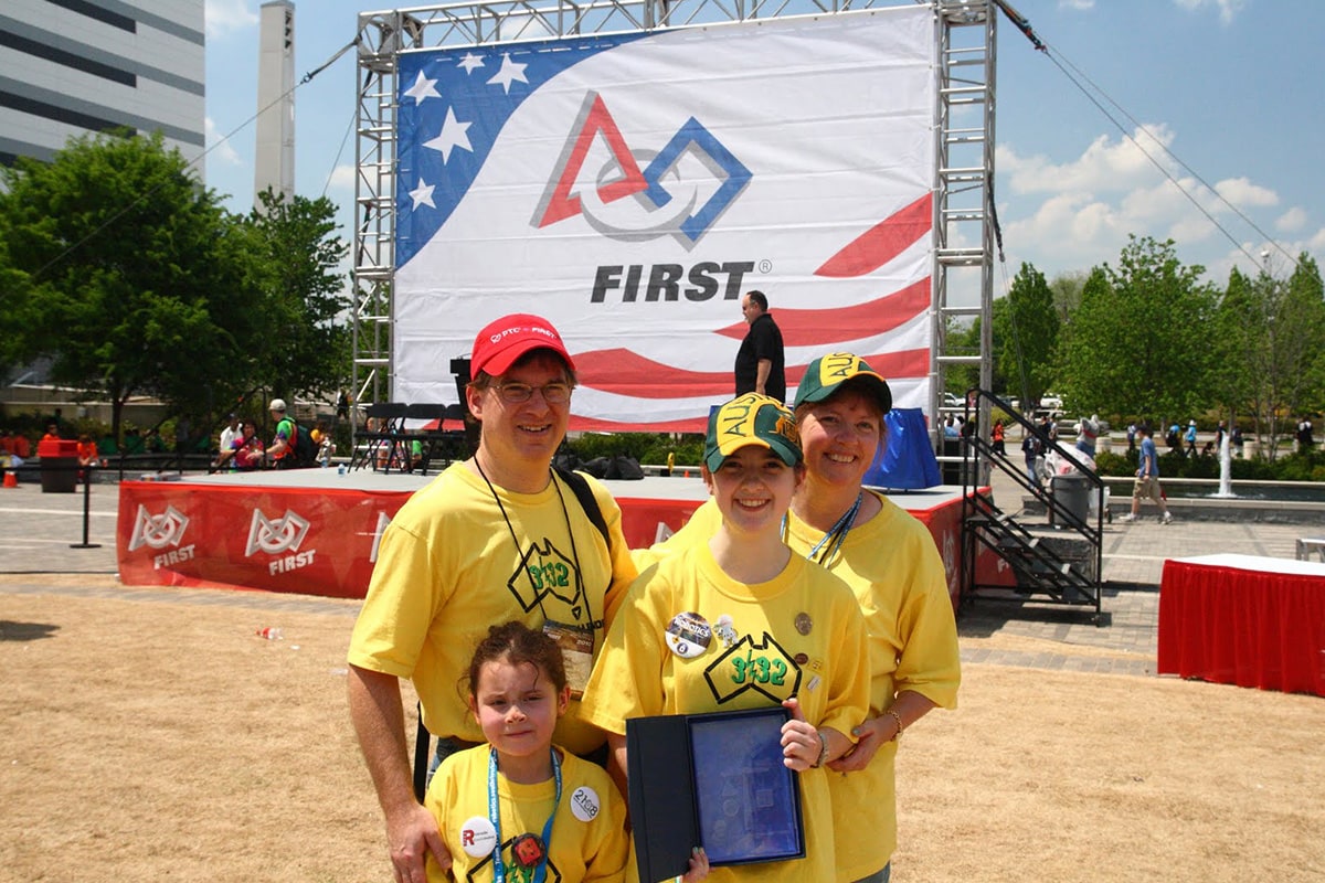 Sarah Heimlich and family pose in front of a large FIRST banner, highlighting early robotics experiences that inspire STEM careers.