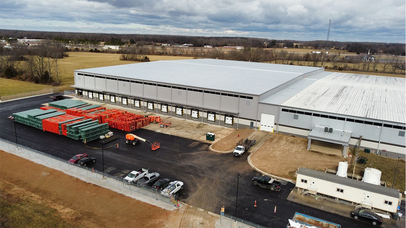 Aerial view of the John Deere Reman facility expansion in Missouri, featuring warehouse buildings, loading docks, parked vehicles, and staged construction materials on site.