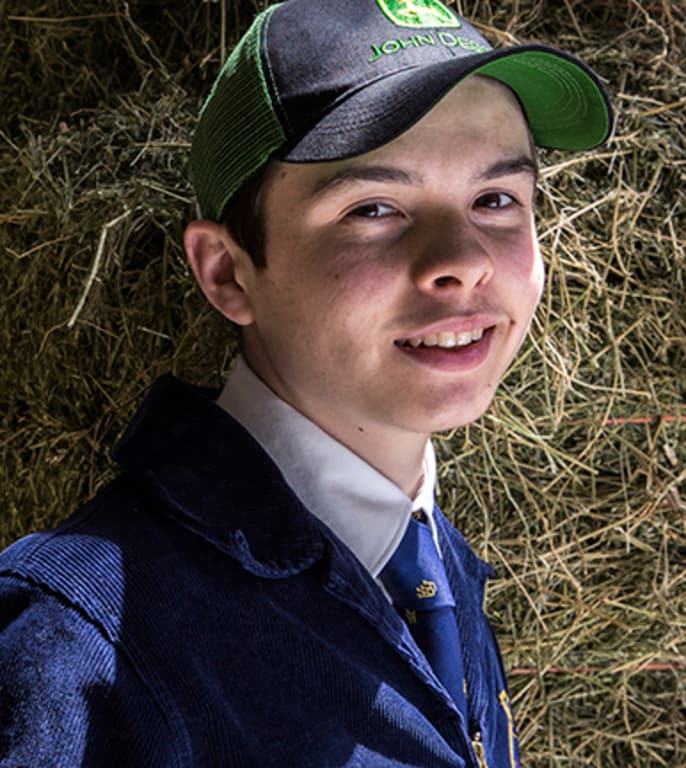 An FFA student wearing a John Deere hat stands in front of a hay bale An FFA student wearing a John Deere hat stands in front of a hay bale