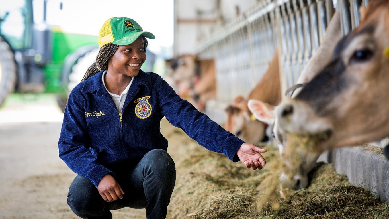 A teenage girl crouches down and reaches her hand out to feeding cows