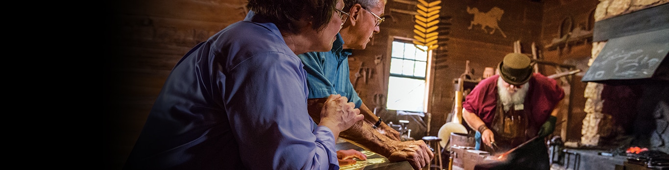 Pareja de ancianos observando al herrero martillar una pieza de metal en el Sitio Histórico de Grand Detour, Illinois, Estados Unidos