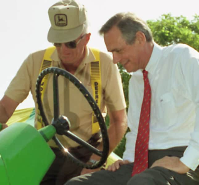 Hans Becherer sitting on a tractor seat with a farmer standing on a step showing him how it operates