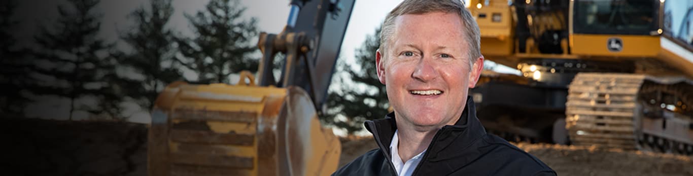 CEO and Chairman John May standing in front of a John Deere excavator at a construction site	