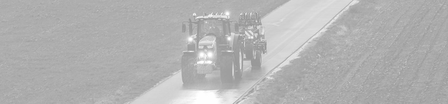 Toned black and white photograph of a 6M Series tractor on a road with its lights on transporting a cultivator on a green background