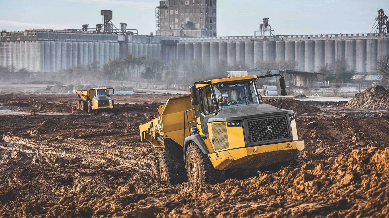 John Deere E-II ADTs in the dirt in front of warehouses