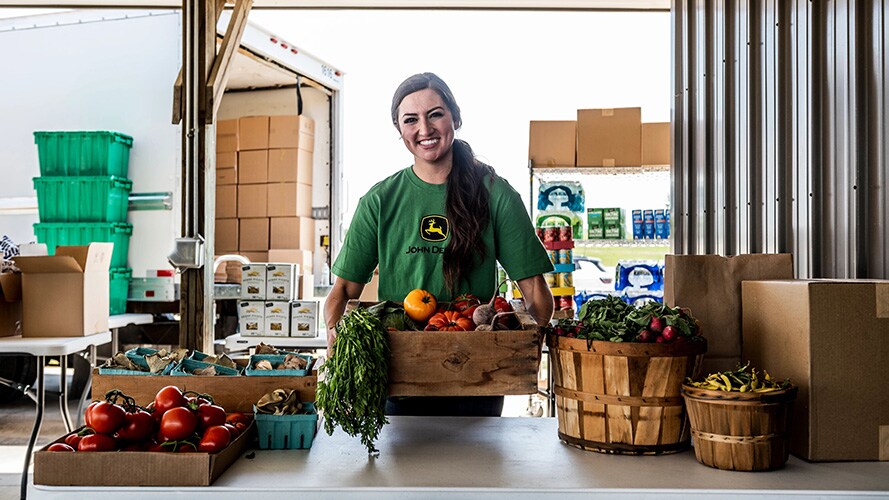 A woman in a John Deere shirt with many baskets of fresh fruit in front of her.