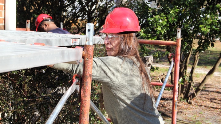 Woman wears hard-hat and safety glasses working near scaffolding.