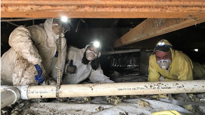 Three people doing clean-up work under a house wearing head lamps and masks, one motions a thumbs up.