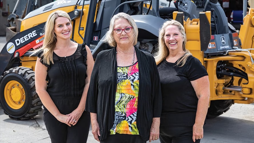 Amanda Matson, Janet Ferguson, and Nikki Worden of Newman Backhoe Service stand in front of a John Deere 310L Backhoe