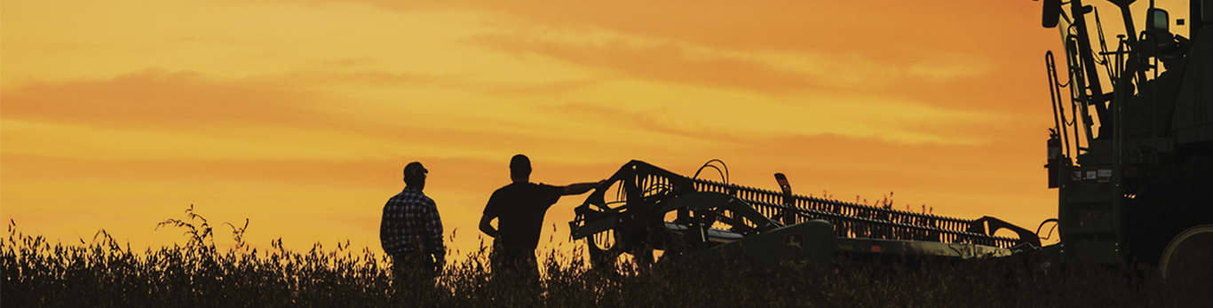 Two farmers standing in a field with a John Deere harvester next to them.