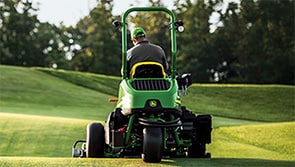 Person operating a turf machine on a golf fairway with trees in the background.