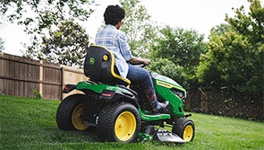 Person operating a riding lawn mower on grass with trees and a fence in the background. 