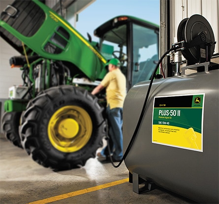 A person fills a John Deere 7230 Row Crop Tractor from a tank of PLUS-50 II Premium Engine Oil SAE 15W-40 inside a garage.
