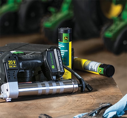 A John Deere 20V Lithium-Ion Battery Powered Grease Gun on a work bench next to two tubes of John Deere Multi-Purpose SD Polyurea Grease, work gloves, and tools.