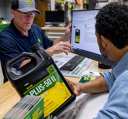 A customer and a John Deere dealer look at a computer screen displaying information about John Deere Plus-50 II Premium Engine Oil SAE 15W-40.