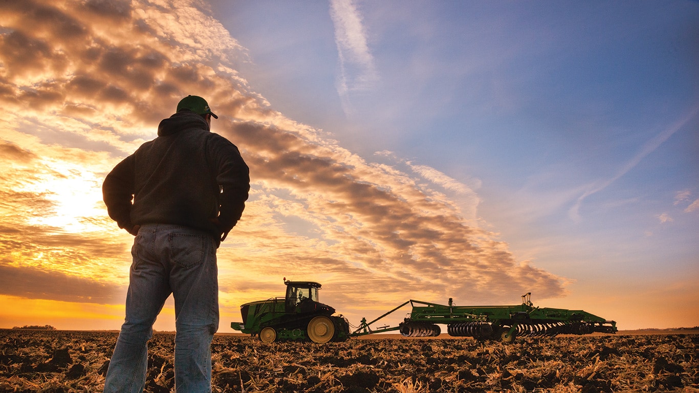 A farmer standing in a harvested field with a John&nbsp;Deere tractor in the distance.