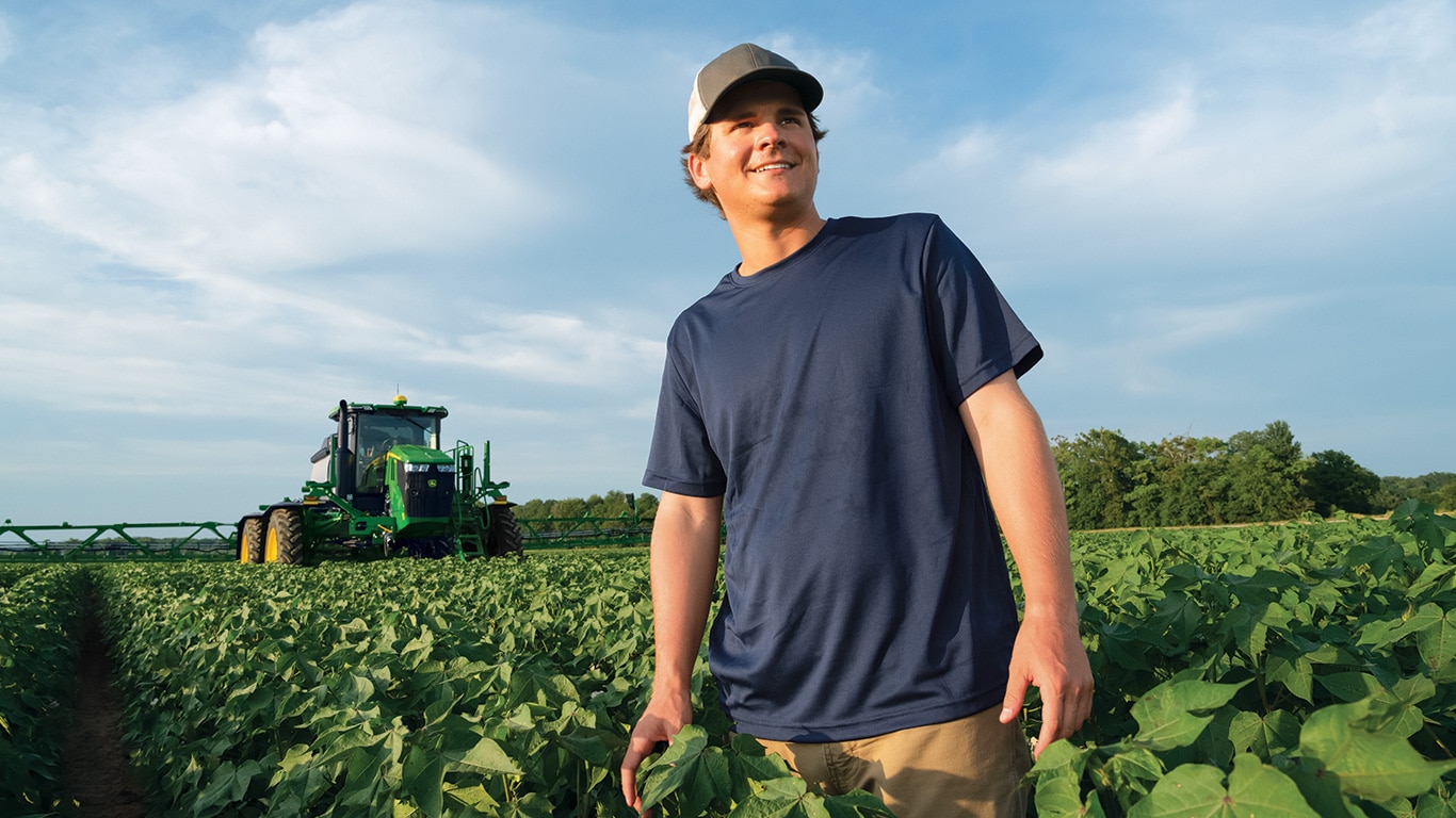 A smiling farmer standing in a soybean field with a John&nbsp;Deere tractor in the distance.