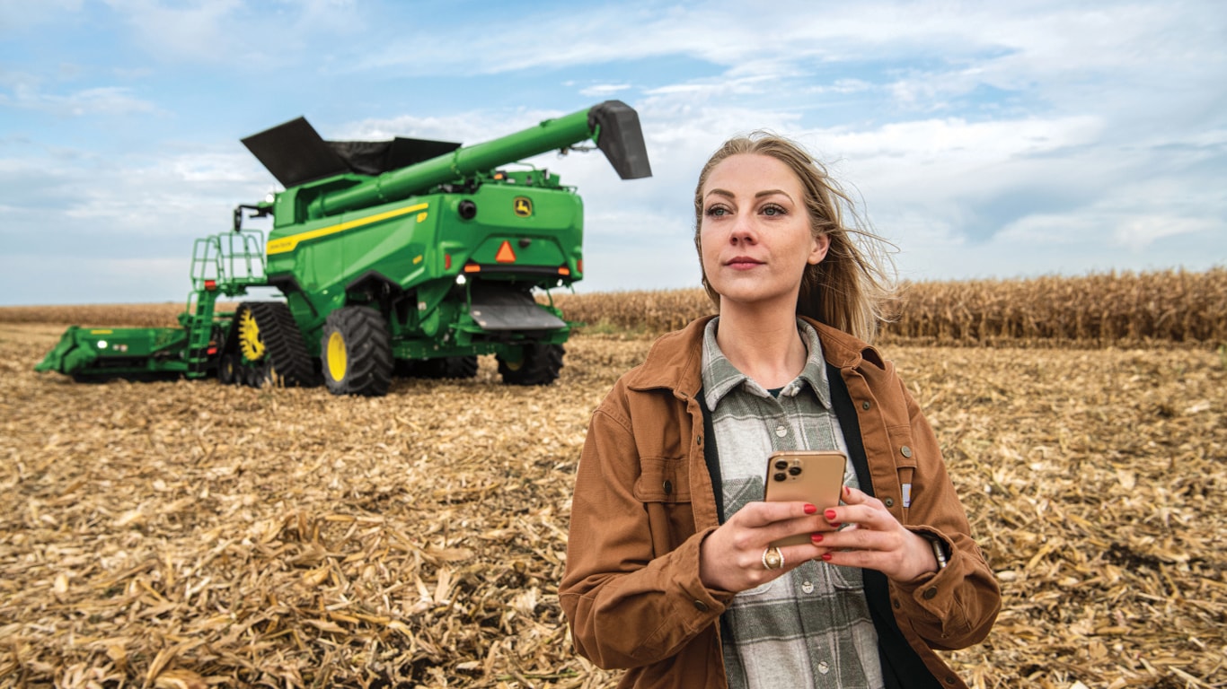 A farmer standing in a partially-harvested cornfield, holding an iPhone in front of a John&nbps;Deere combine.