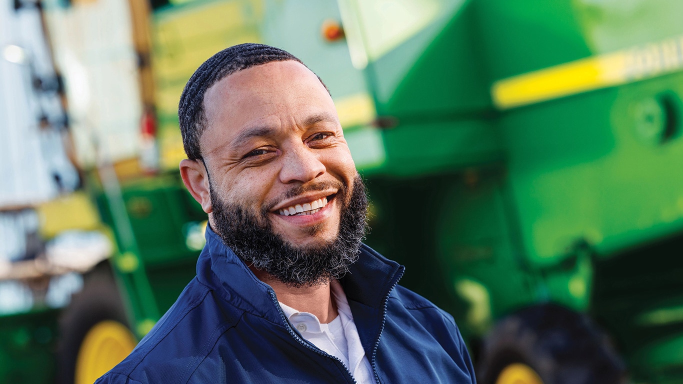 Close-up of a smiling farmer standing in front of a John&nbsp;Deere harvester.