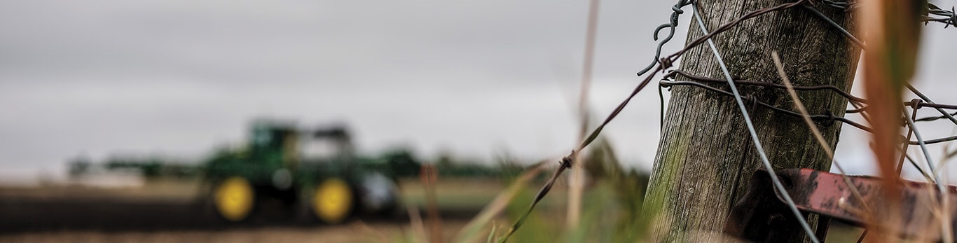 Close-up of a wooden fence post wrapped in barbed wire with a green tractor blurred in the background.